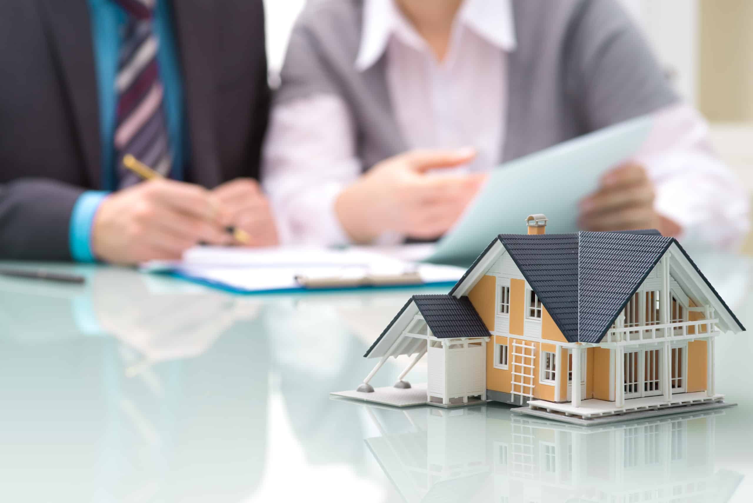 Image of home architectural model on a table with two people sitting at the table one signing a document.